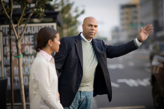 Mid-adult man hailing a taxi while standing next to a woman on the side of an inner city street.