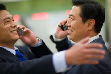 Two smiling businessmen congratulating each other while talking on their cell phones.