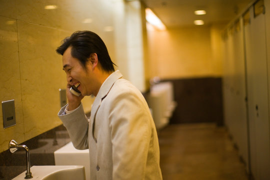 Mid-adult man laughing while talking on his cell phone in a public bathroom.