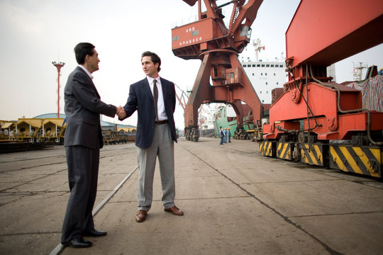Young adult businessman shaking hands with a mid-adult male colleague at shipping yard.