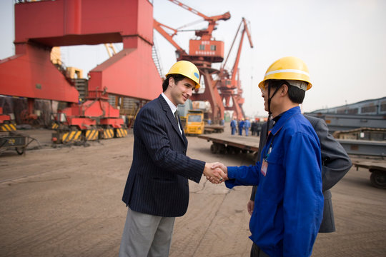 Young adult businessman shaking hands with a mid-adult foreman at a shipping yard.