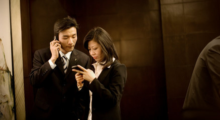 Young adult businessman on a cellphone while a mid-adult female colleague texts in a board room.