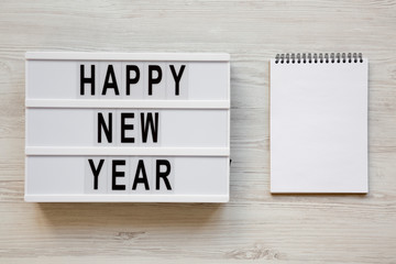 Modern board with text 'Happy new year' and blank notepad over white wooden background, top view. Overhead, flat lay, from above. 2019