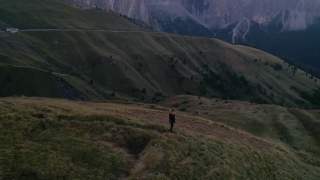 Woman standing on a hill, C4k aerial, descending drone shot, of a girl hiking on a hill, in front of Dolomiti mountains, at Sella pass, on a sunny, summer morning, in Dolomites, Tyrol, Italy