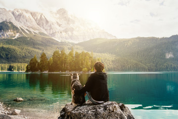 Mensch umarmt Hund auf einem Felsen mit Blick auf einen wunderschönen See mit Bergen und Bäumen