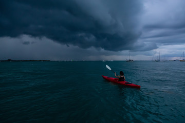 Adventurous girl on a red kayak is kayaking towards a thunderstorm during a dramatic sunset. Taken...