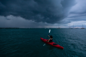Adventurous girl on a red kayak is kayaking towards a thunderstorm during a dramatic sunset. Taken...