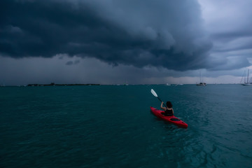 Adventurous girl on a red kayak is kayaking towards a thunderstorm during a dramatic sunset. Taken in Key West, Florida Keys, United States.