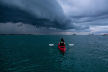 Adventurous girl on a red kayak is kayaking towards a thunderstorm during a dramatic sunset. Taken...