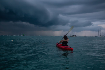 Adventurous girl on a red kayak is kayaking towards a thunderstorm during a dramatic sunset. Taken...