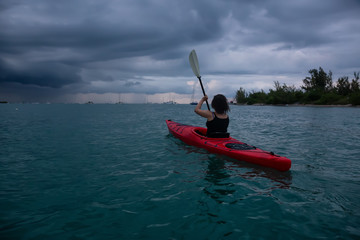 Adventurous girl on a red kayak is kayaking towards a thunder storm during a dramatic sunset. Taken...