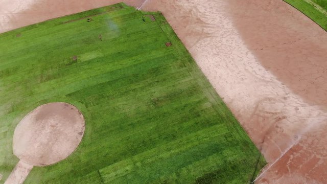 Slow motion drone shot of a baseball diamond's sprinkler watering the turf under the afternoon sun.