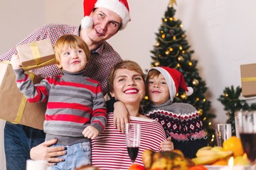 Happy family parents with children in Santa Claus hats stand at the Christmas table with gifts. Joyful family holiday