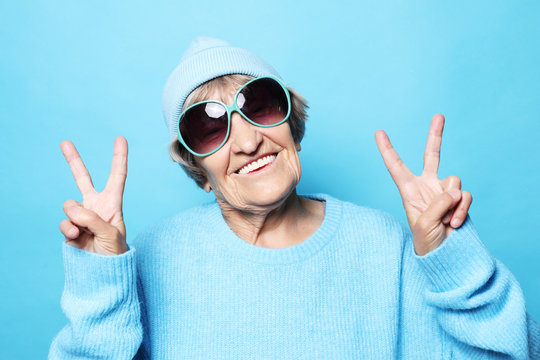 Lifestyle, emotion and people concept: Funny old lady wearing blue sweater, hat and sunglasses showing victory sign.