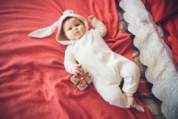 Smiling cute baby child in rabbit costume lying with gift on red bed with gift near Christmas tree