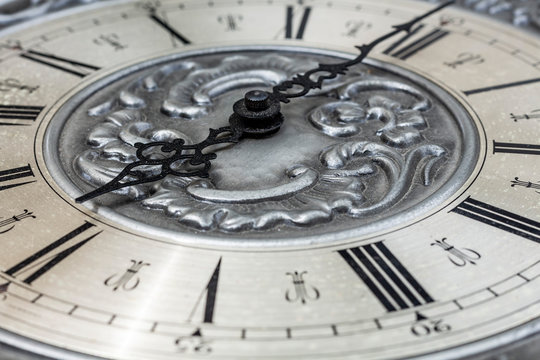 Old vintage clock with metal dial and floral embossed pattern close-up. Black clock hands indicating a time of 6 hours. Beautiful vintage patterned dial with Latin numerals