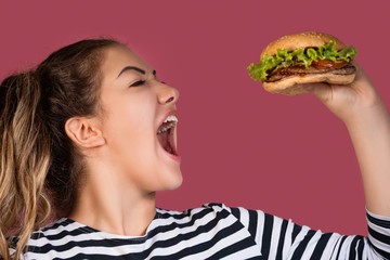 Hungry cool girl in striped t-shirt eating hamburger over pink background