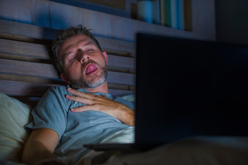 man alone in bed playing cybersex using laptop computer watching porn sex movie late at night with...