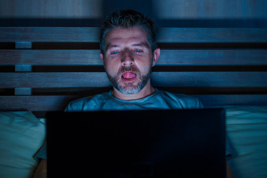 man alone in bed playing cybersex using laptop computer watching porn sex movie late at night with lascivious pervert face expression in internet pornographic sexual content