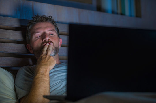 man alone in bed playing cybersex using laptop computer watching porn sex movie late at night with lascivious pervert face expression in internet pornographic sexual content