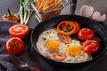Fried eggs with bacon and tomatoes on an old cast-iron pan with french fries on a gray table. Close-up