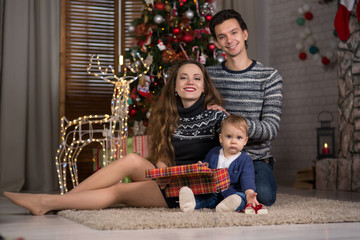 family sits with the baby sitting near the Christmas tree,