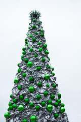 Fragment of decorative silver color metallic material crumpled christmas tree with green balls on light background with copy space for text. Festive winter holidays decor.
