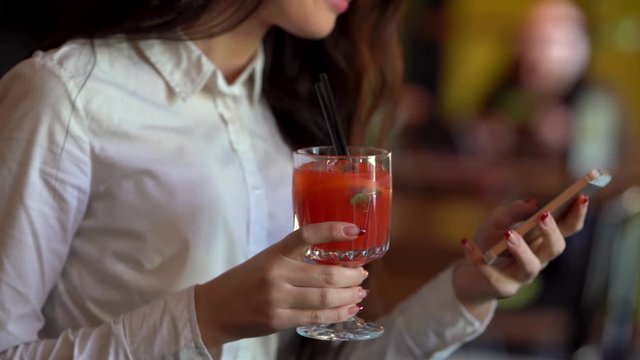 The darkhair girl in white blouse in one hand is holding red cocktail and in the other one her cellphone, looking at cellphone and smiling.