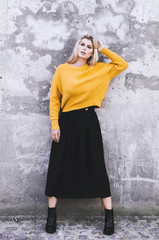 A girl in a yellow sweater, wearing a black skirt and boots in the background of a concrete wall. Clothes in style oversize