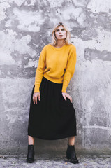 A girl in a yellow sweater, wearing a black skirt and boots in the background of a concrete wall. Clothes in style oversize