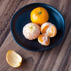 Black plate of satsuma oranges on a rustic wood table, two oranges peeled, one sectioned, and peels on the table