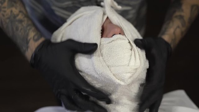 Tattoed hands in black gloves of a barberman tightly wrapping face of a male customer in a white towel after shaving. Close-up concept