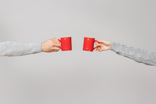 Close up cropped of woman, man two hands horizontal holding red cups of tea, clinking isolated on grey wall background. Friends leisure, healthy lifestyle, ill cold season concept. Mock up copy space.