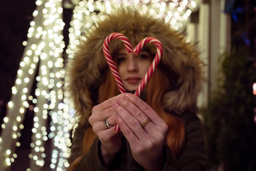 Red haired girl with candy heart on the street. On the background garland. Girl in the defocus. Valentines day concept