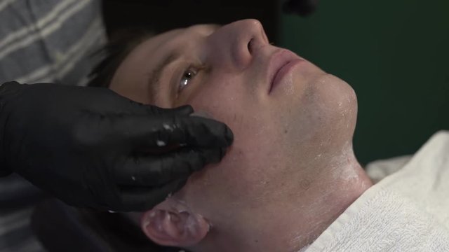 Tattooed hands in black gloves of a barberman taking off/removing the white towel off male customer's face. Barberman rubbing client's face with special cube-shape aroma oil. Close-up concept