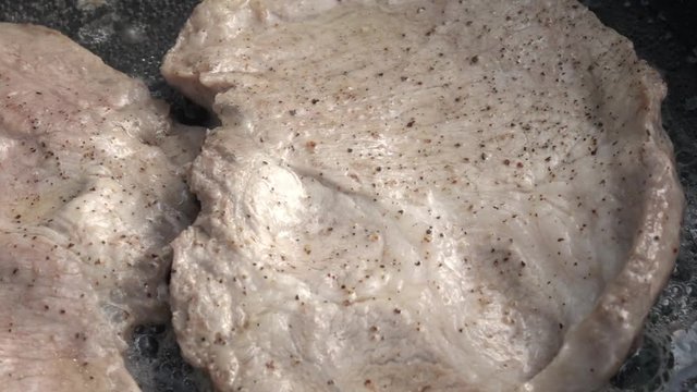 Slow motion of sizzling pork steaks on dark gray fry pan close up. Appetizing natural food background with amazing texture. Mouthwatering home cooking view. High speed shooting without post processing