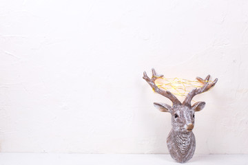 Decorative head of New Year reindeer with fairy light on its antlers on white textured background.