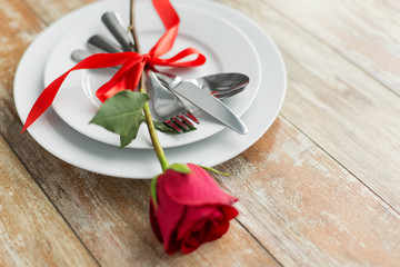 valentines day and romantic dinner concept - close up of red rose flower on set of dishes with cutlery on wooden table