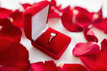 jewelry, proposal and romantic concept - diamond ring in red velvet gift box on bed sheet and rose...