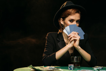 attractive girl in jacket and hat covering face with poker cards and looking at camera in casino
