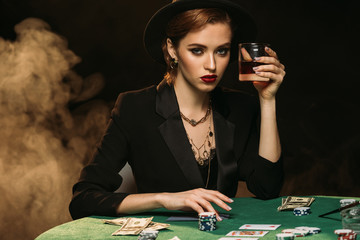 attractive girl in jacket and hat holding glass of whiskey at poker table in casino and looking at...