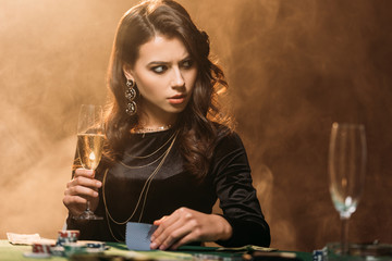 irritated attractive girl holding glass of champagne at poker table in casino and looking away