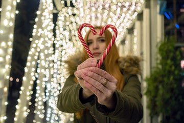 Red haired girl with candy heart on the street. On the background garland. Girl in the defocus. Valentines day concept