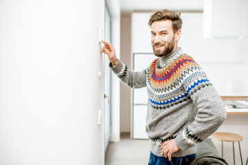 Man in sweater adjusting room temperature with electronic thermostat at home