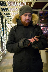 bearded man with a phone in winter clothes on the street. garland on the background