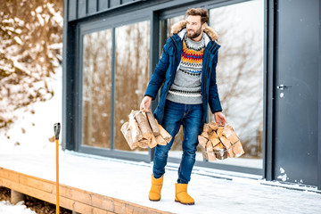 Handsome man in winter clothes carrying firewoods on the terrace near the modern house in the mountains