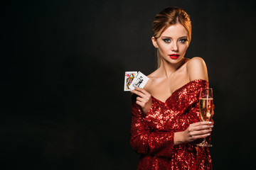 attractive girl in red shiny dress holding joker and queen of hearts cards isolated on black,...