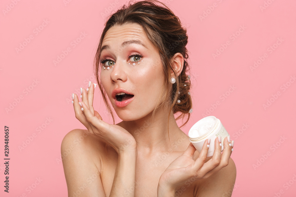 Sticker Pretty young woman posing isolated over pink wall background take care of her skin with cream.