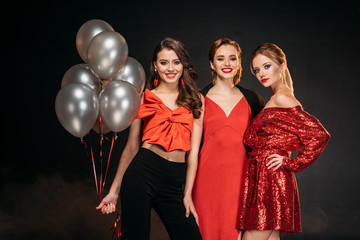 smiling attractive girls in red clothes holding bundle of grey balloons isolated on black