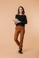 Photo of charming woman 20s holding book while standing, isolated over beige background